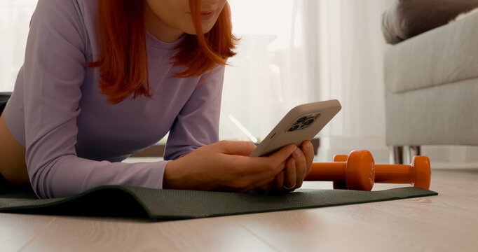 A woman relaxes on a gym mat in her living room, taking a break post-exercise. She scrolls through her smartphone, enjoying a moment of downtime after her workout.