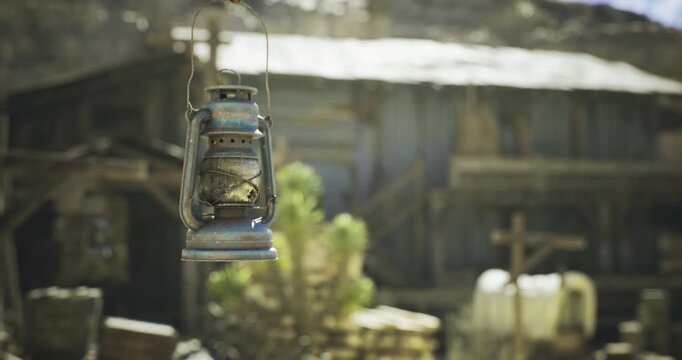 A rustic lantern dangles from a hook, surrounded by wooden structures in a historic western settlement. The sun shines brightly, casting shadows and enhancing the old world charm.