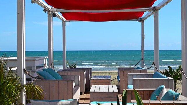 Summer beach lounge area with wooden furniture and red canopy overlooking turquoise sea waves motion.