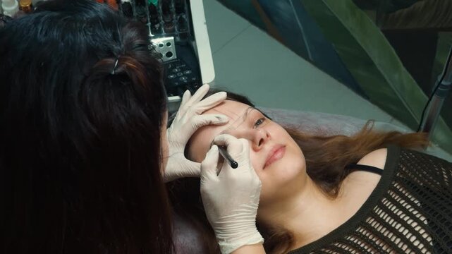 Woman undergoing an eyebrow microblading service, lying on a procedure couch as a specialist meticulously draws permanent makeup hairstrokes, preparing her brows for cosmetic tattooing