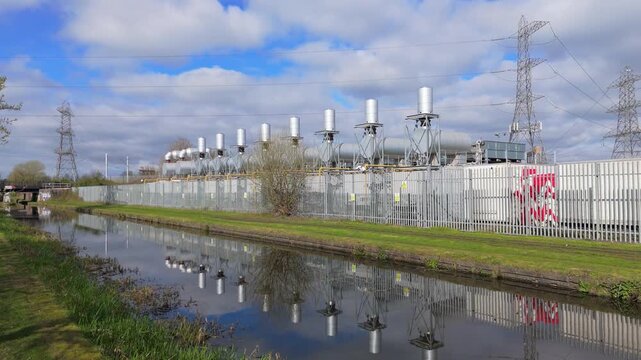 A stationary view of a row of transformers in containers and cooling units above with reflections rippling in the canal water.