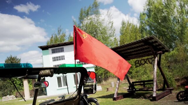 Close up view of red flag with hammer and sickle symbol waving in wind from military utility vehicle, parked outdoors with residential building and trees in background, sunny day celebration