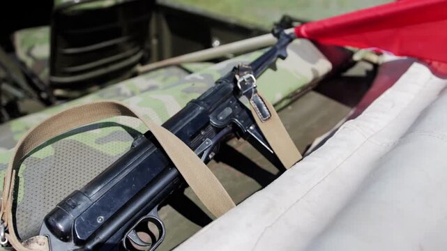 Close-up shot of classic submachine gun with metal and wood components, canvas sling, positioned on mottled green and brown military camouflage material, part of large red flag unfurled in distance, s