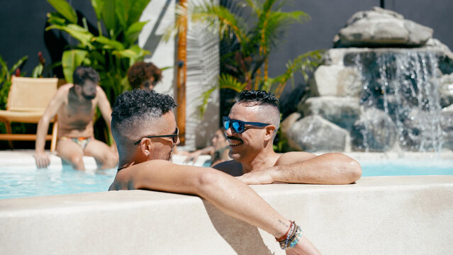 Group of diverse male friends enjoying summer vacation in a tropical swimming pool with waterfall, laughing and socializing