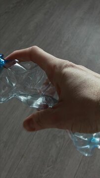 Human hand crushing clear plastic water bottle. Plastic material crumples, showing wrinkles and folds. Simple gray wooden floor background. Depicts waste reduction action and recycling.