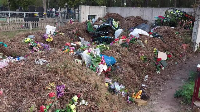 View of accumulated organic debris, including withered leaves and branches, mixed with colorful artificial floral arrangements and trash bags at cemetery refuse site outdoors.