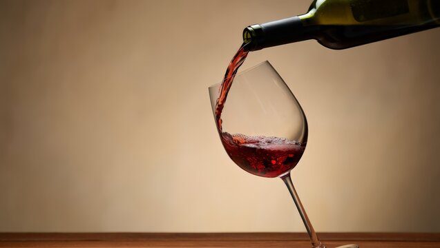 A bottle of red wine is being poured into a glass on a wooden table against a beige background, capturing a moment of luxury and relaxation.