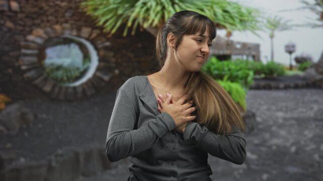 Young woman clutching chest with both hands to heart, eyes closed in a pained smile, standing beside a round stone wall and lush plants on a street; quiet gratitude.