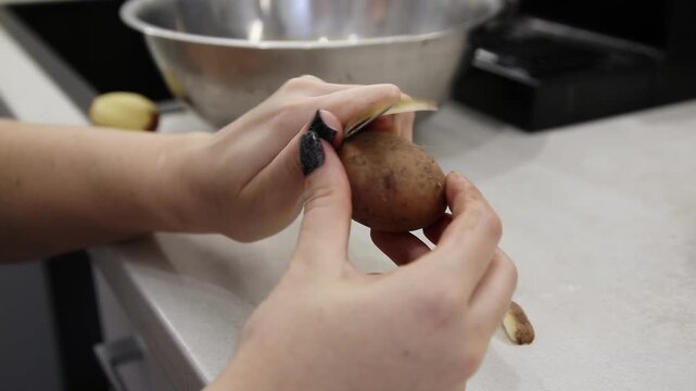 Detailed view of woman hands peeling brown skin of raw organic potato with kitchen peeler tool. Food preparation activity for healthy cooking in modern kitchen environment.