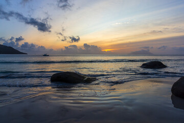 Obraz premium Golden sunset paints pastel skies above a rocky beach as soft waves wash the sand and a lone boat drifts on the calm ocean horizon, creating a peaceful, contemplative seascape mood