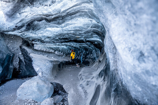 View of a lone figure in a vibrant yellow jacket stands amidst the cool, blue depths of an ice cave, the light filtering through the frozen formations, Sveitarfelagio Hornafjordur, Iceland.