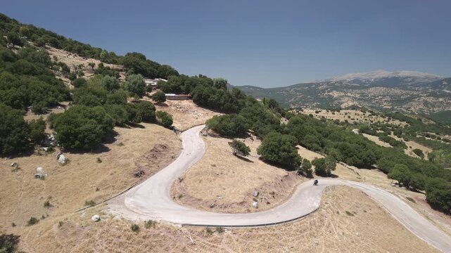 Static shot of a motorcycle riding through a curved mountain pass road in Achaea, Peloponnese, Greece on a sunny day.