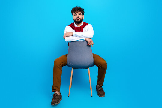 Confident young man sitting backwards on a chair in casual fashion portrait against a bright blue studio background