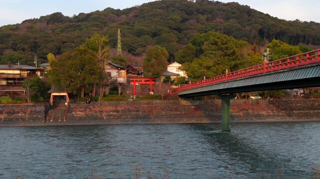 Static shot of the Uji River and the vibrant red Asagiri Bridge, with the traditional Uji Shrine visible in the background against a lush green hill in Uji, Japan.
