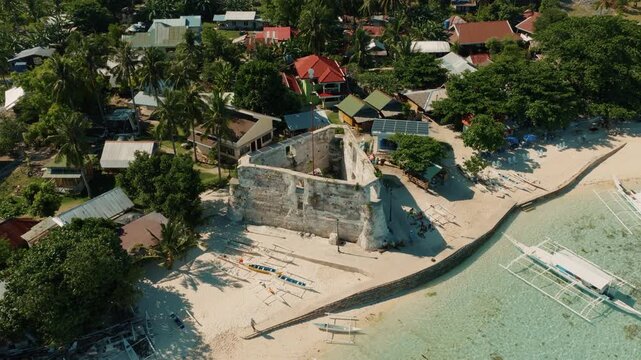 Old stone watchtower ruins beside tropical beach with outrigger boats and village houses Pamilacan Island Bohol, aerial orbit