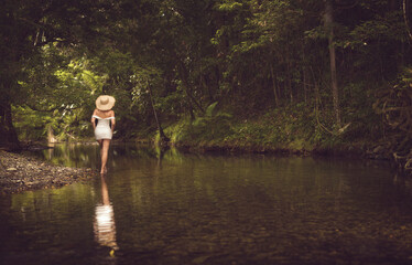 Fototapeta premium an elegant woman in a white dress and sun hat, together with serene reflective landscapes surrounded by lush green forest and soft natural light.