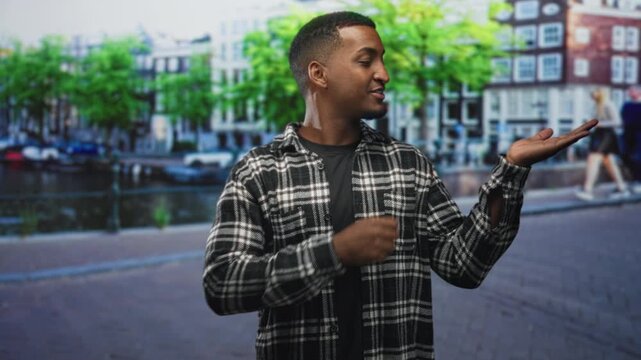 Man in plaid shirt smiling, pointing with finger and showing open hand on street beside a canal and row houses, gesturing to palm; friendly welcome.