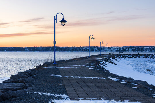 Winding riprapped pier with lampposts seen during a late winter blue hour sunrise with the St. Lawrence river and the south shore in the background, Neuville, Quebec, Canada