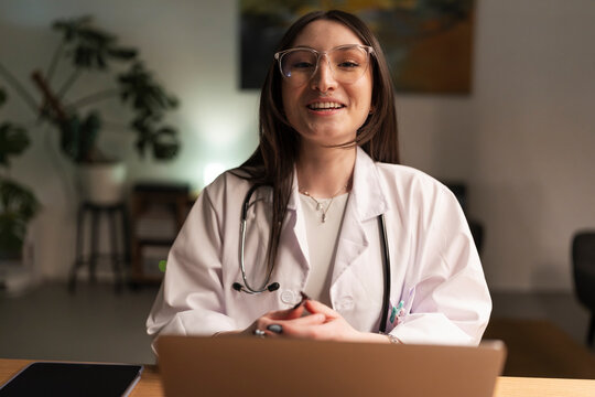A smiling medical professional in a lab coat and stethoscope is seen at a desk with a laptop, engaging in a virtual consultation or online work.