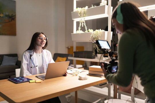 A doctor in a white coat and stethoscope speaks to a camera operator filming her presentation.
