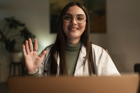 A smiling female doctor wearing glasses and a white coat waves hello during a video call, offering a friendly greeting.