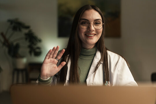 A smiling female doctor in a white coat waves hello during a video call, offering a friendly greeting from her office.