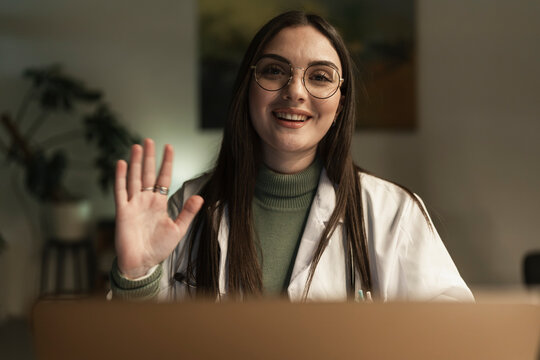 A smiling female doctor in a white coat and glasses waves hello during a video consultation, offering a friendly greeting.