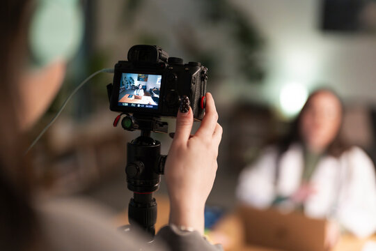 A person films another person sitting at a table with a laptop and tablet, capturing content for a video.