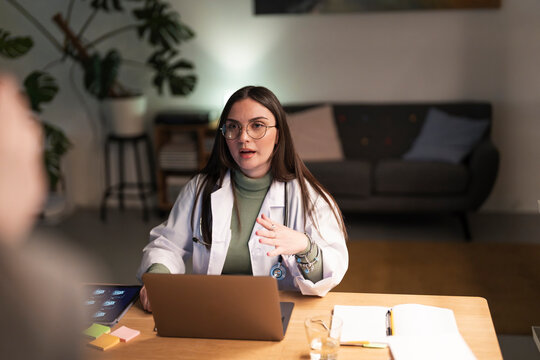 A female doctor in a white coat and glasses is speaking while gesturing with her hands, sitting at a desk with a laptop and medical notes.