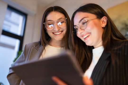 Two young women in business attire smile while looking at a tablet together, sharing an idea or a moment of discovery.