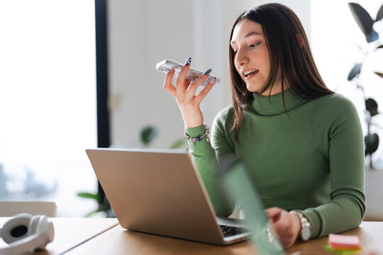 A young woman is working on her laptop and speaking on her phone. She is sitting at a desk with headphones nearby.