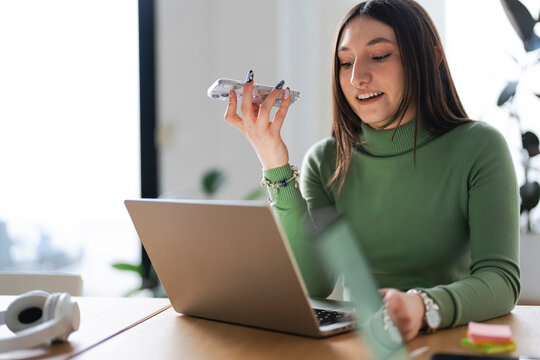A young woman is working on her laptop and speaking into her phone.