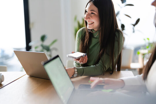 Two women collaborate at a desk with laptops and a smartphone, engaged in discussion and work in a bright, modern office setting.