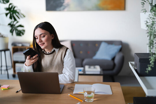 A young woman works on her laptop and uses a calculator while sitting at a desk in a home office.