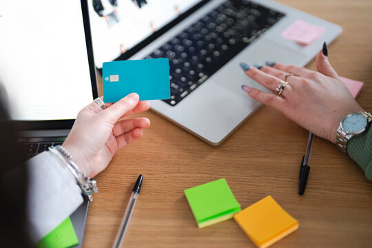 A person holds a debit card while shopping online on a laptop, with colorful sticky notes and pens on the desk.