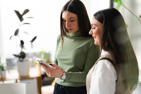 Two women are looking at a mobile phone together, one is smiling while the other is focused on the screen.
