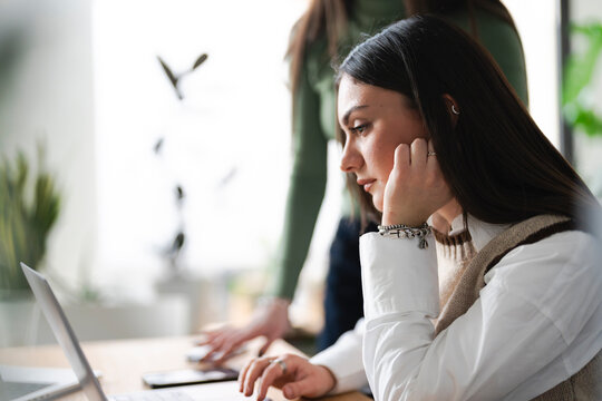 Two women collaborate on a project, one focused on a laptop screen while the other gestures towards a tablet, suggesting teamwork and shared ideas.
