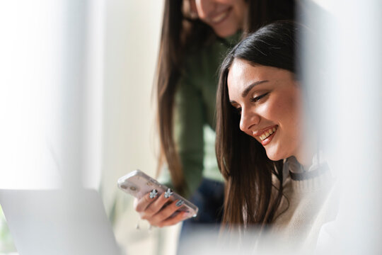 Two young women share a moment of laughter while looking at a mobile phone and a laptop, suggesting collaboration or shared digital content.
