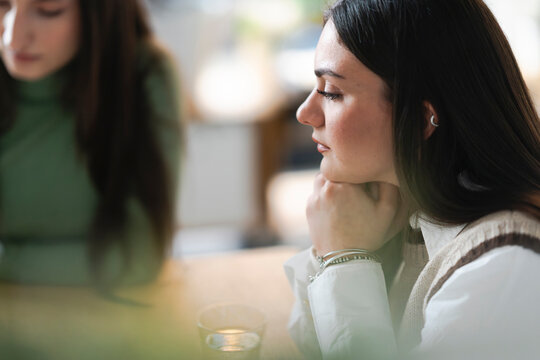 A young woman rests her chin on her hands, deep in thought while sitting at a table with a drink.