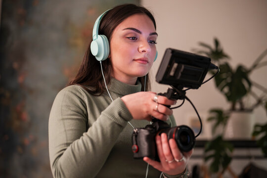 A young woman wearing headphones holds a camera with a monitor attached, focusing on her work.