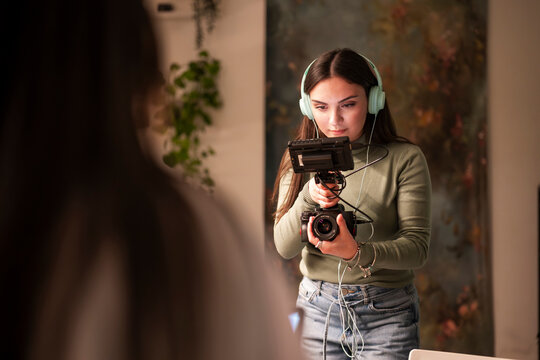 A young woman wearing headphones operates a professional camera and monitor, capturing footage in a studio setting.