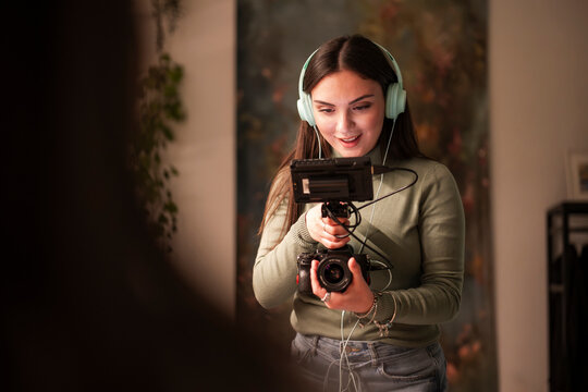 A young woman wearing headphones operates a video camera and a digital camera, focused on her work.