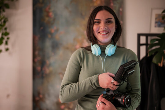 A young woman with headphones around her neck holds a camera and a recording device, ready to capture content.