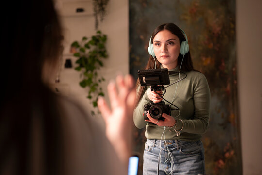 A woman wearing headphones holds a camera and monitor, filming someone in front of her.