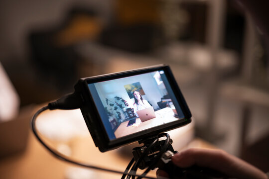 A person films a woman working on a laptop in a studio setting, captured through a camera monitor.