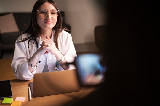 A young female doctor in a white coat and glasses sits at a desk with a laptop, tablet, and sticky notes, ready to consult with a patient.