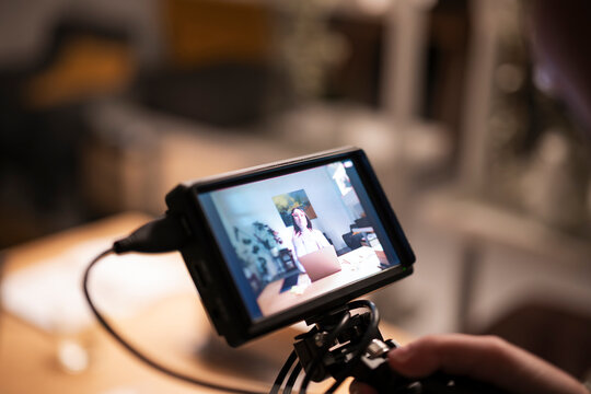 A person's hand operates a camera monitor displaying a woman working on a laptop in a home office setting, capturing content for online distribution.