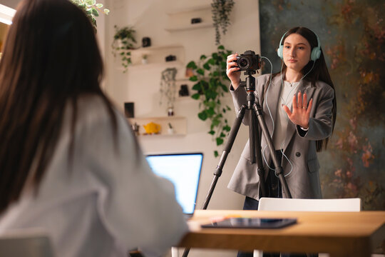 A young woman wearing headphones holds a camera on a tripod, directing a recording session in a home studio.