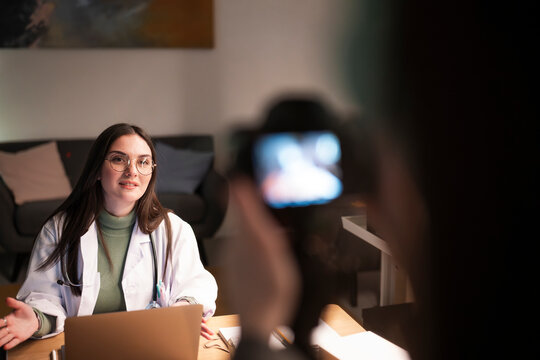A doctor in a white coat speaks to a camera while sitting at a desk with a laptop and stethoscope.