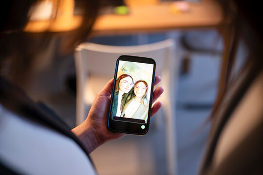 A person holds a smartphone displaying a selfie of two smiling young women. They are posing for a photo together.